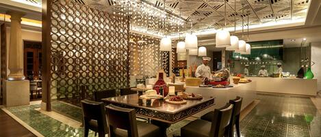 Un ristorante con un design geometrico del soffitto, un tavolo centrale circondato da sedie e uno chef che cucina al bancone.