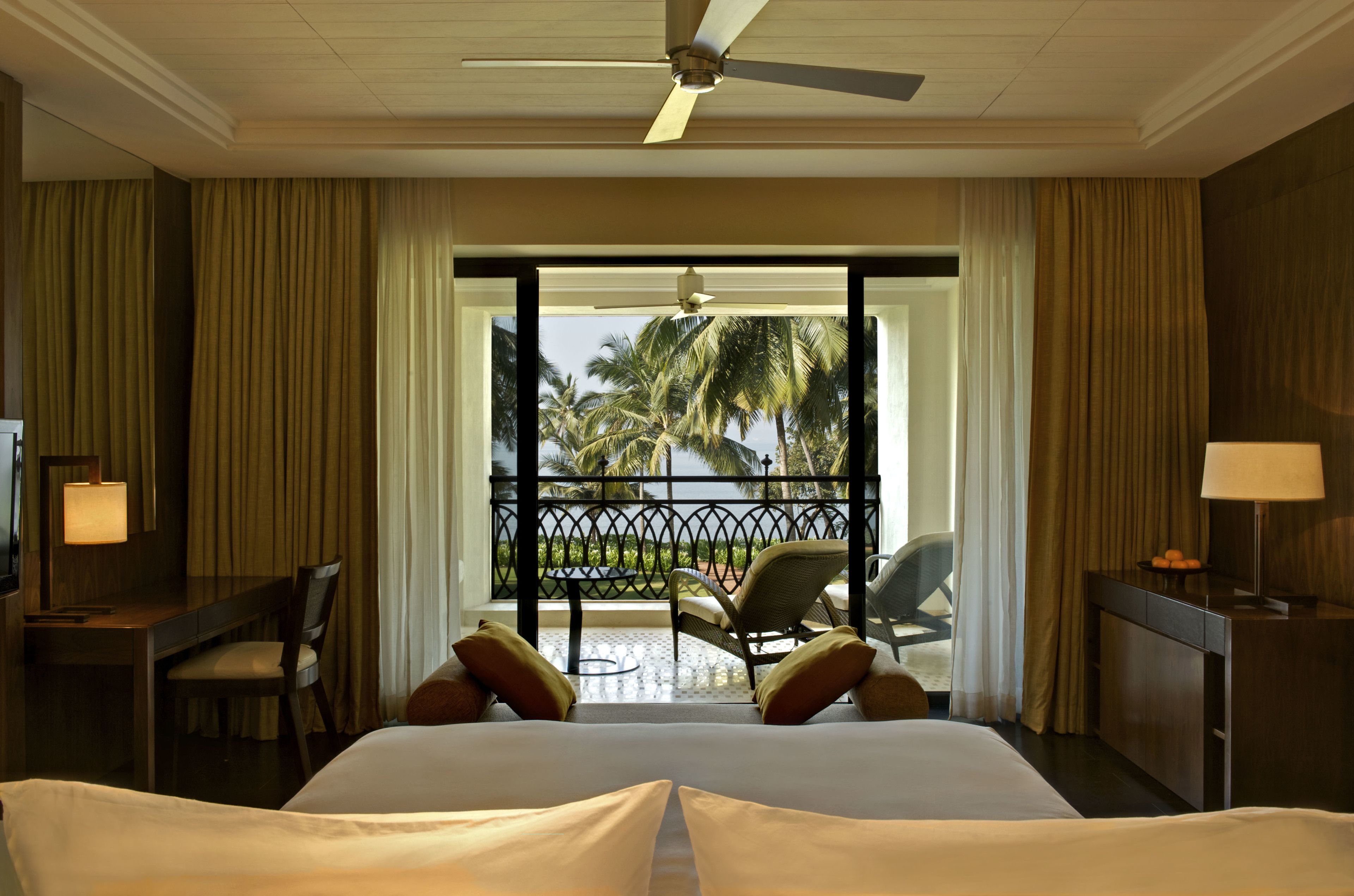 A hotel room with a bed, a desk with a lamp, a chair, and a view of palm trees through a balcony door.