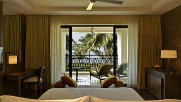A hotel room with a bed, a desk with a lamp, a chair, and a view of palm trees through a balcony door.