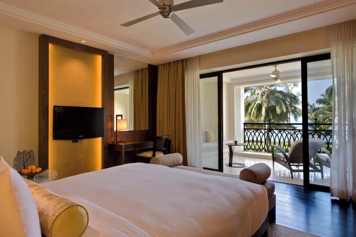 Habitación de hotel con una cama grande, un escritorio con silla, televisor y un balcón con vistas a palmeras.