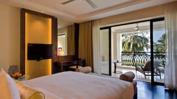 A hotel room with a large bed, a desk with a chair, a television, and a balcony with a view of palm trees.