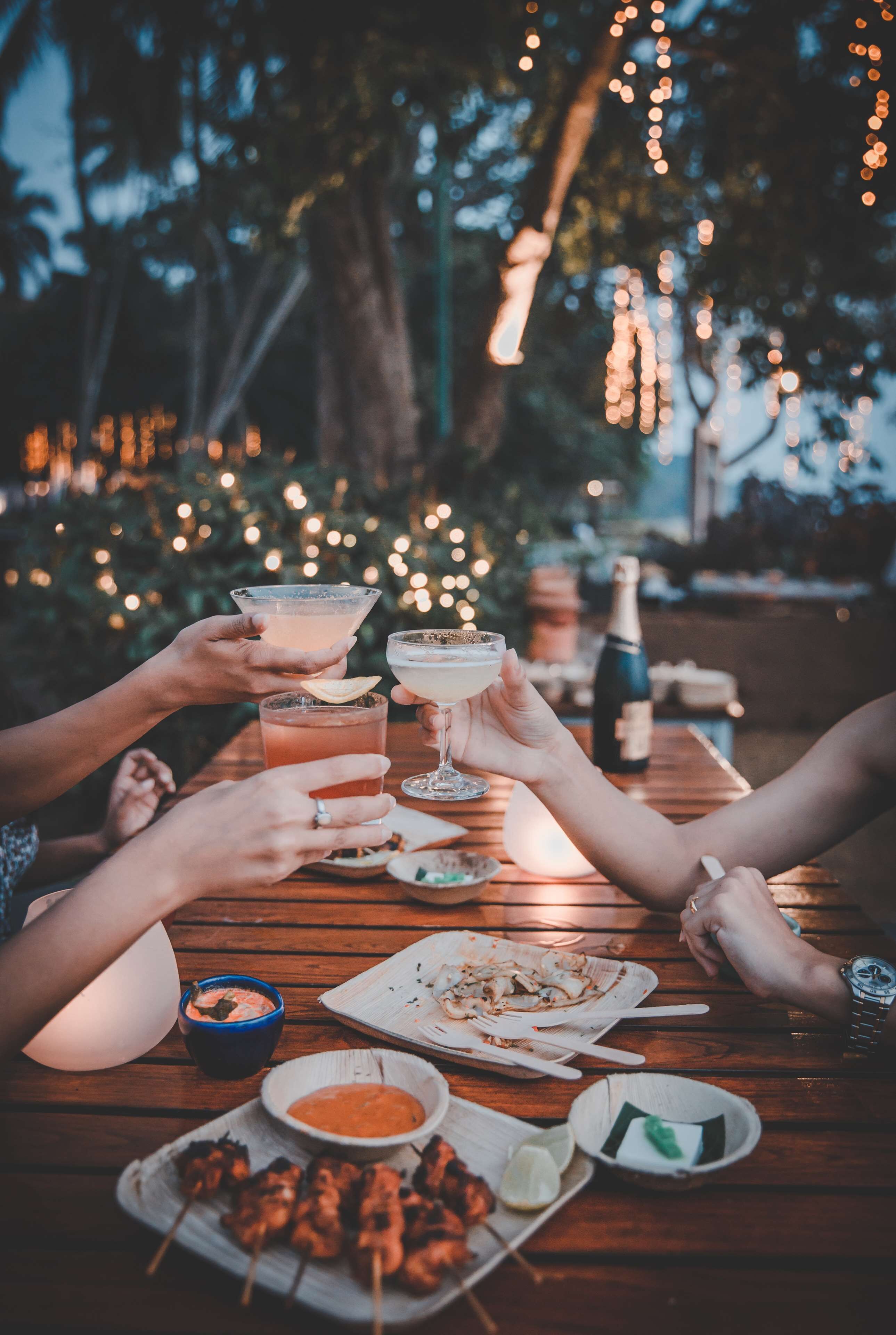 A group of people toasting with drinks at a wooden table outdoors, with string lights in the background.