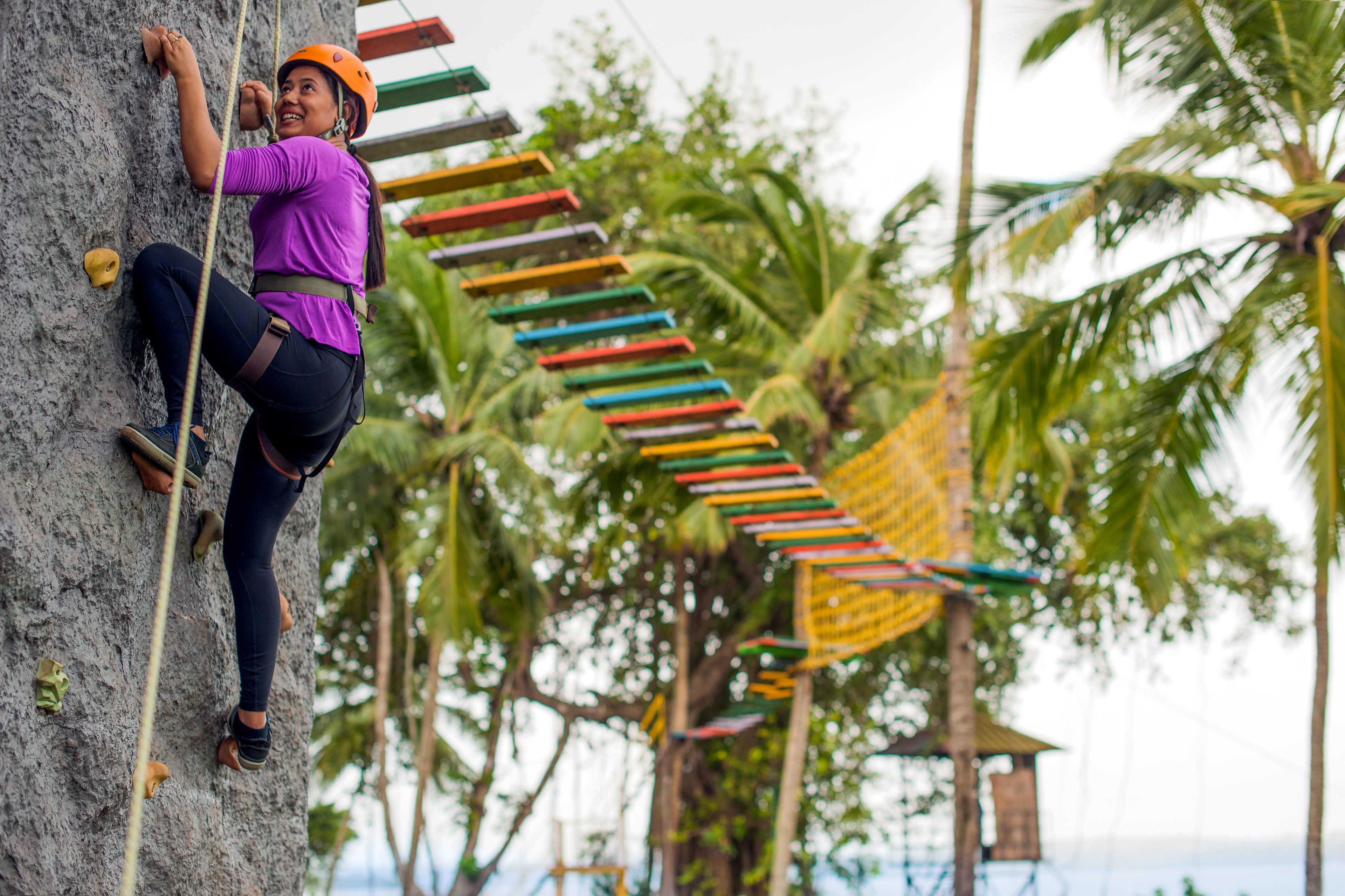 Una persona escalando una pared de roca con barandillas de seguridad coloridas y palmeras al fondo.
