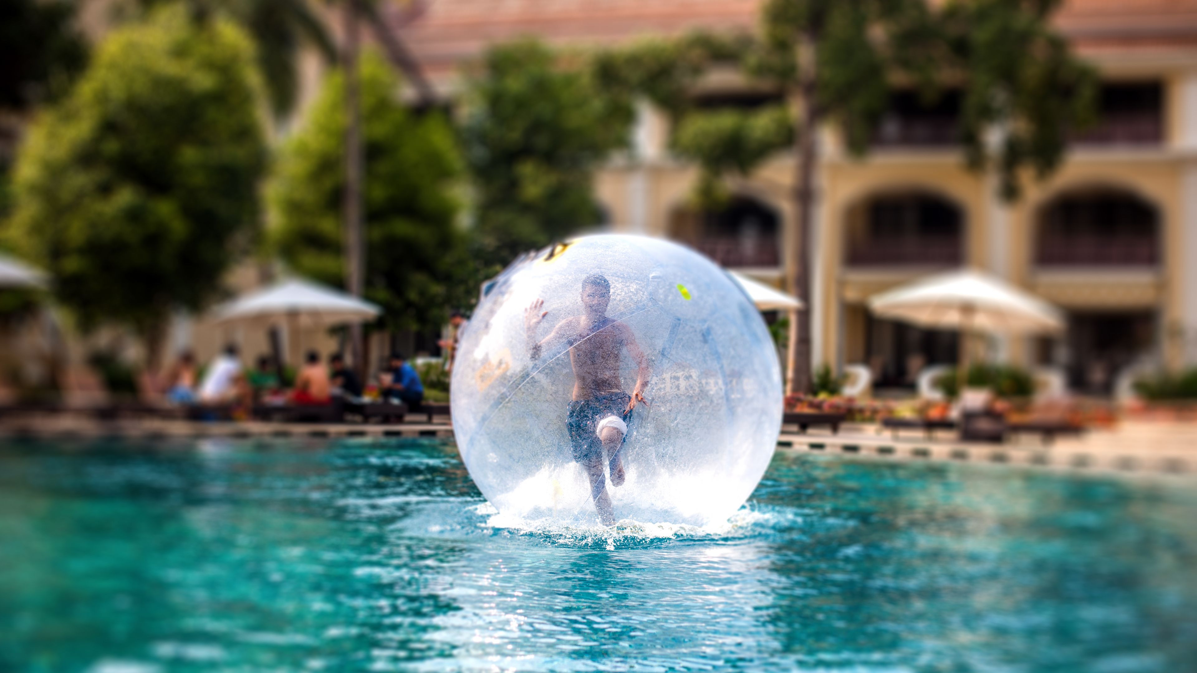 A person inside a transparent sphere in a pool, with a building and lounge area in the background.