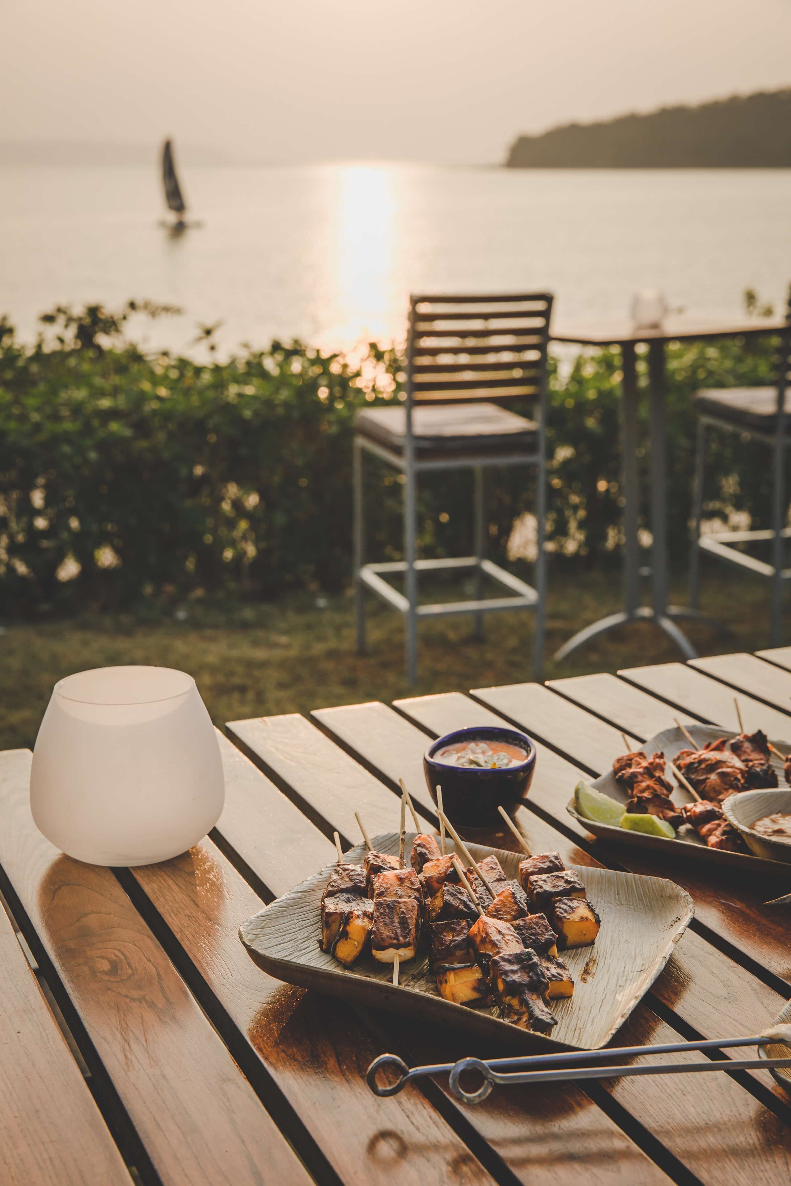 A wooden table with skewers of grilled meat, a bowl of sauce, and a glass of water.