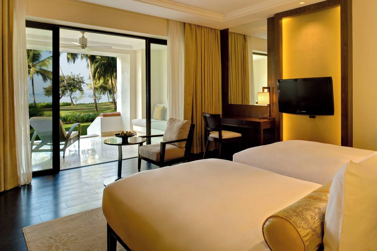 Habitación de hotel con dos camas, un escritorio, una silla, televisión y vistas a palmeras y un cuerpo de agua.