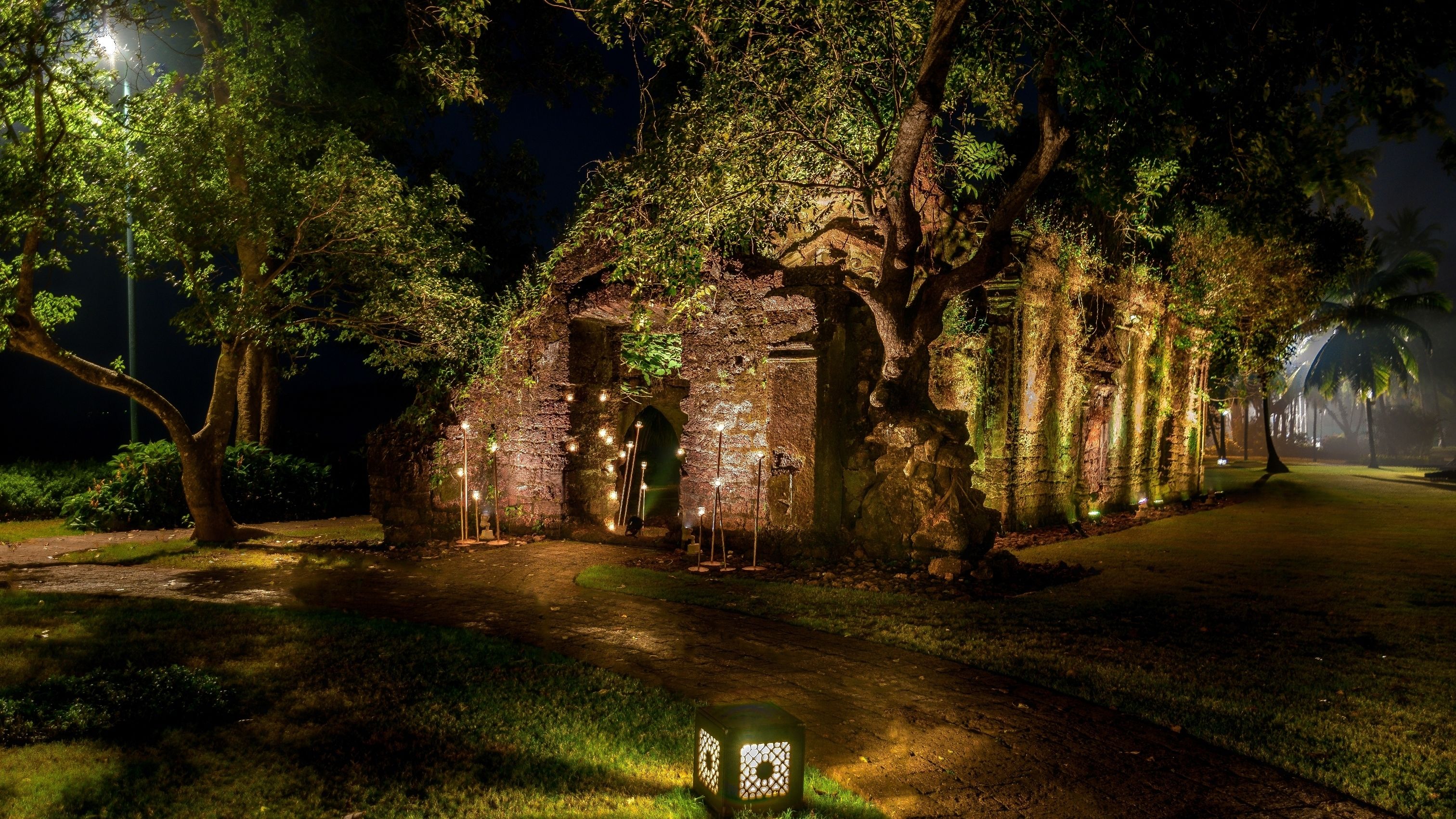 Una estructura de piedra con un arco central, rodeada de árboles e iluminada por varias fuentes de luz.