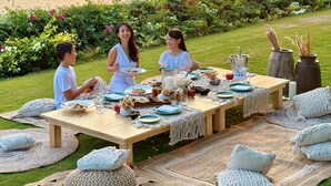 A picnic setup with a table and chairs on grass, overlooking a body of water at sunset.