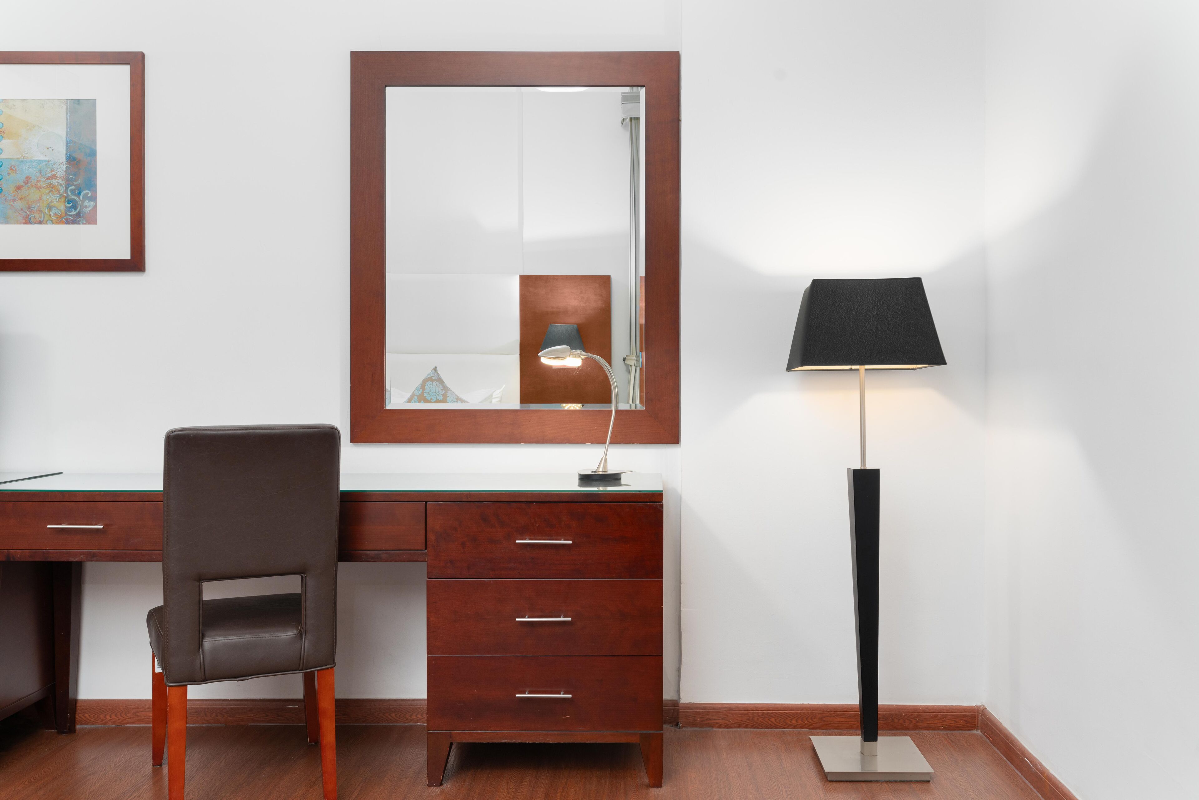 Un escritorio de madera con una silla, un espejo y una lámpara de pie en una habitación con paredes blancas y suelo de madera.