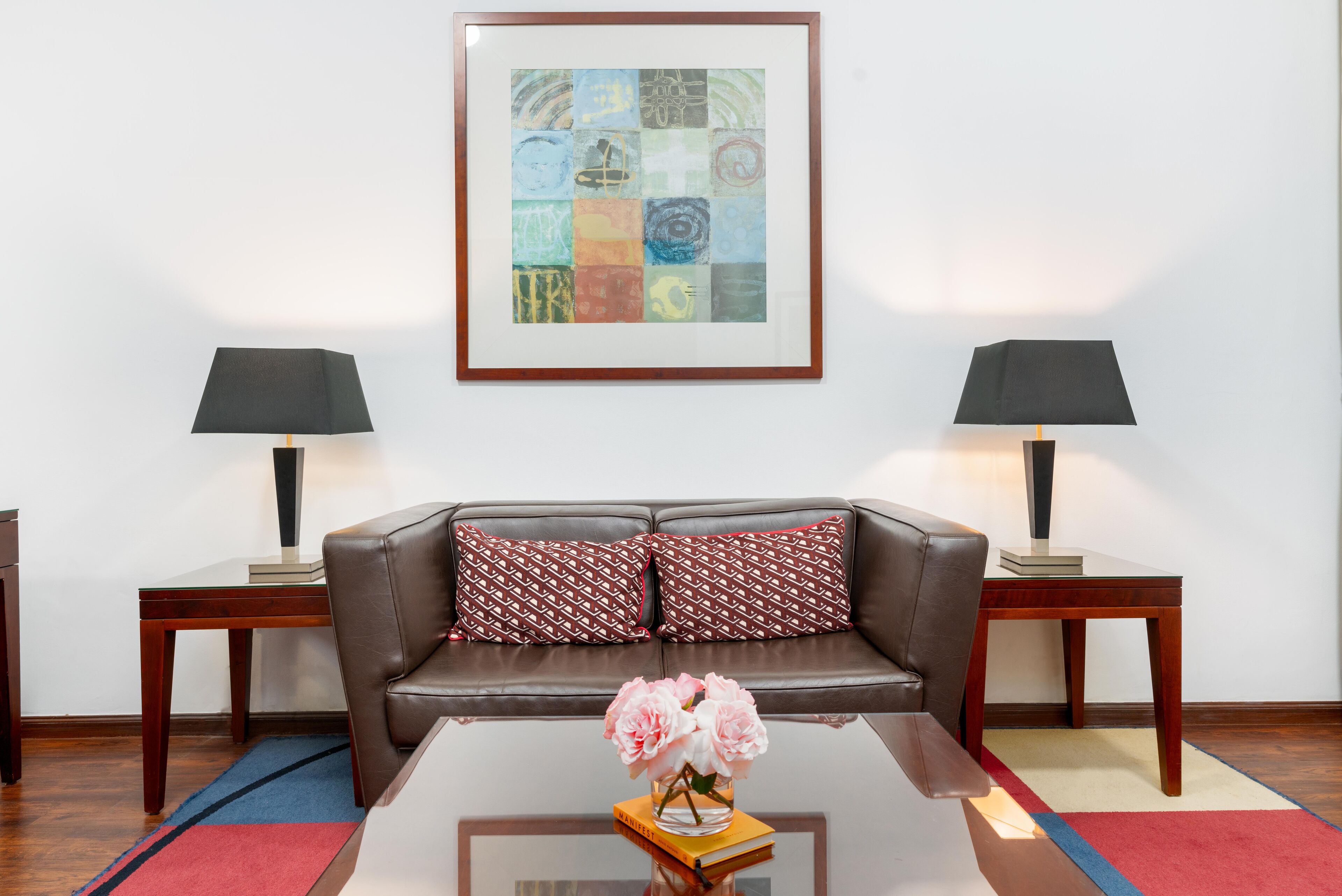 Una sala de estar con un sofá de cuero marrón, dos mesitas de noche con lámparas, una obra de arte abstracta enmarcada y un jarrón con flores sobre una mesa de centro.