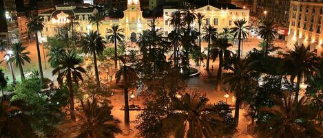A well-lit square with palm trees and benches, surrounded by historic buildings with illuminated facades.