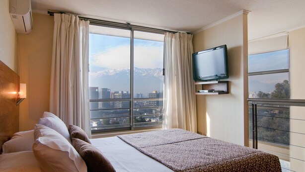 A hotel room with a bed, a TV mounted on the wall, and a large window offering a city and mountain view. - Park Plaza Apart Hotel (Santiago)