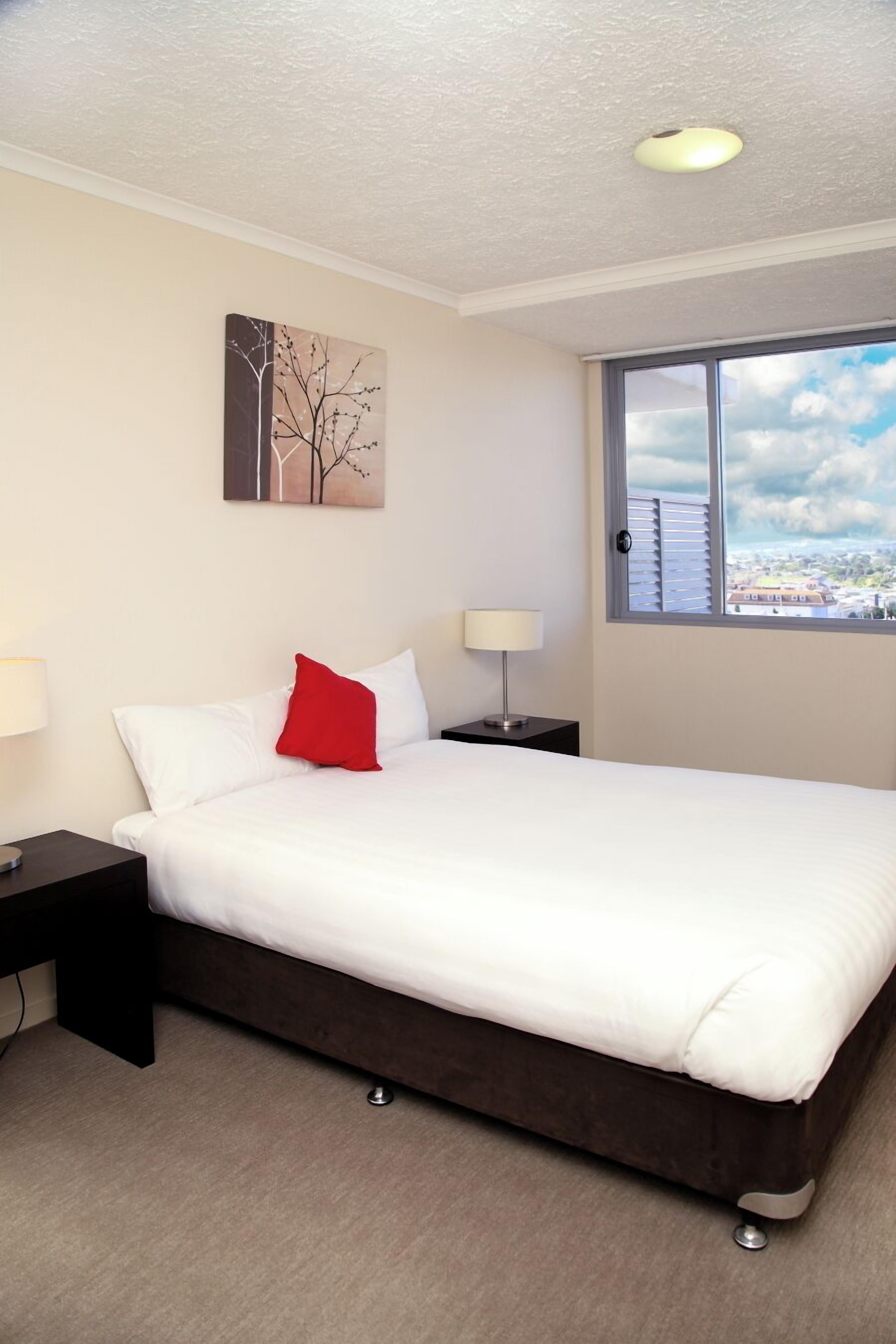 A hotel room with a bed, a nightstand, a lamp, a window with a city view, and a painting on the wall.