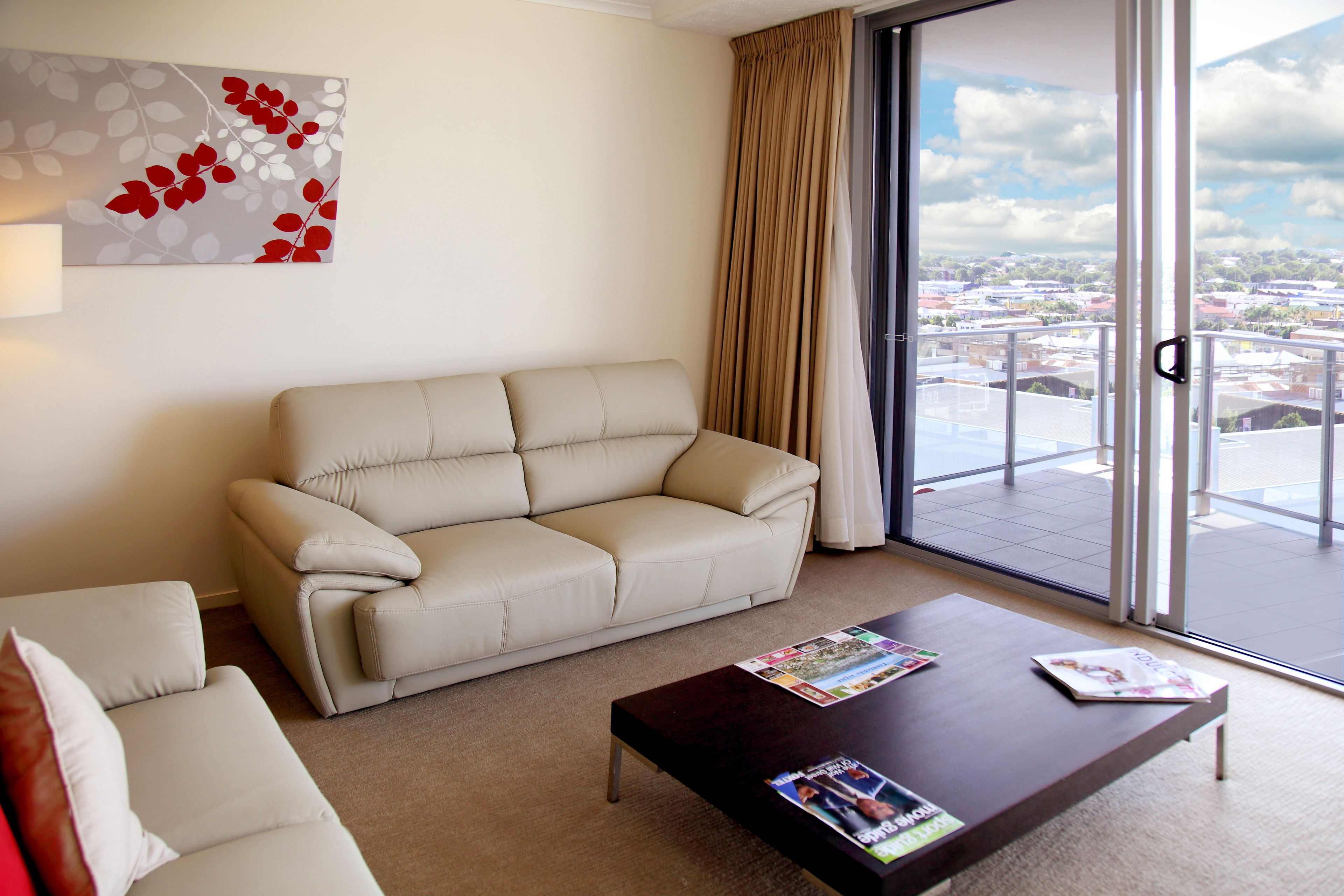 Una sala de estar moderna con un sofá color beige, una mesa de centro con revistas, un ventanal con vistas a un paisaje urbano y una obra de arte abstracto colgada en la pared.