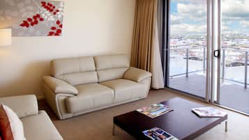 Una sala de estar moderna con un sofá color beige, una mesa de centro con revistas, un ventanal con vistas a un paisaje urbano y una obra de arte abstracto colgada en la pared.