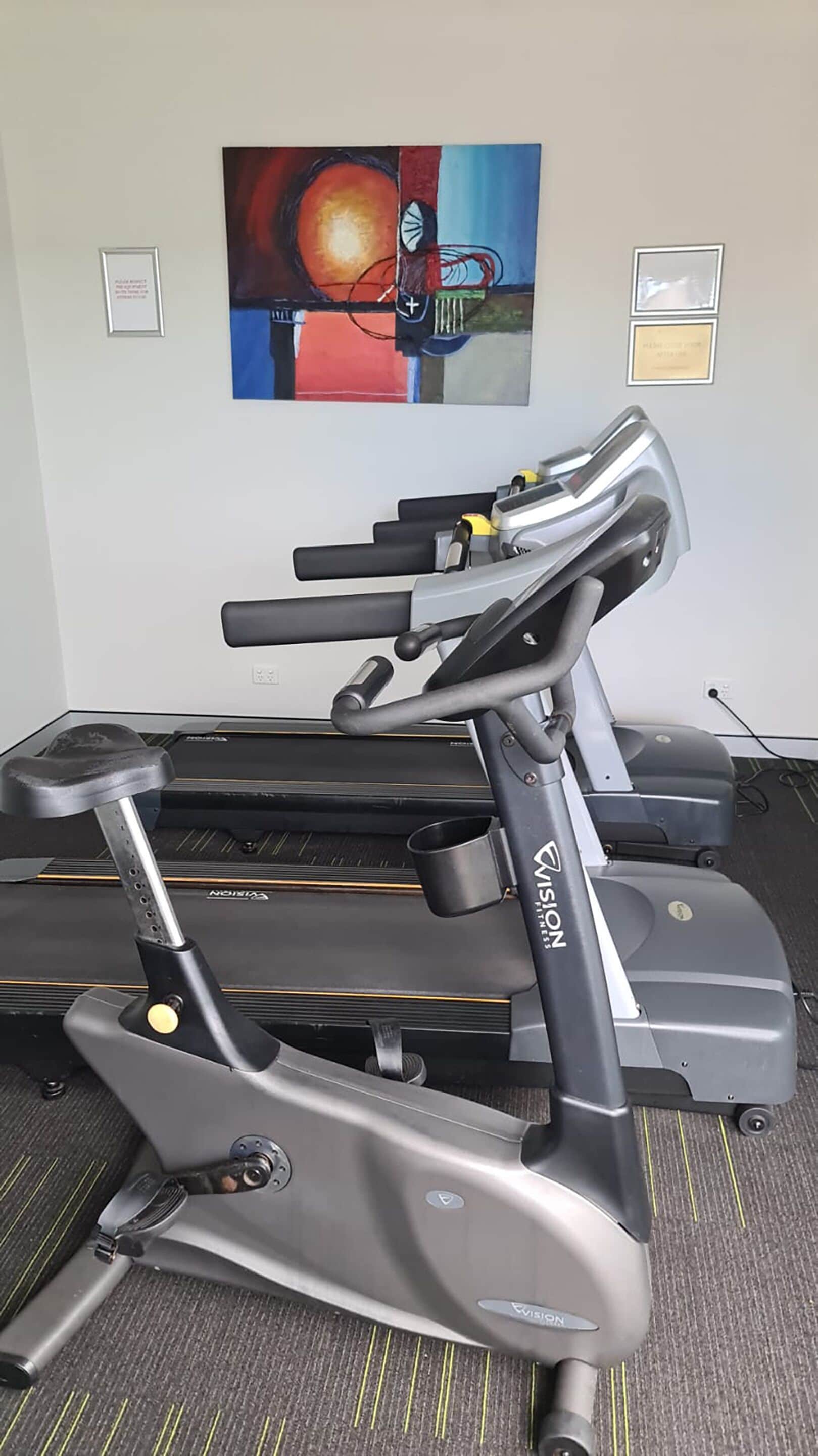 A stationary bike with a control panel and a display screen, placed in a room with a carpeted floor and a painting on the wall.