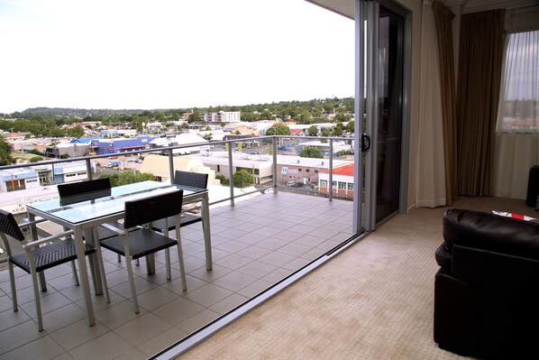 Un balcón con puerta de vidrio, una mesa de comedor con sillas y vista a un pueblo con edificios y árboles.