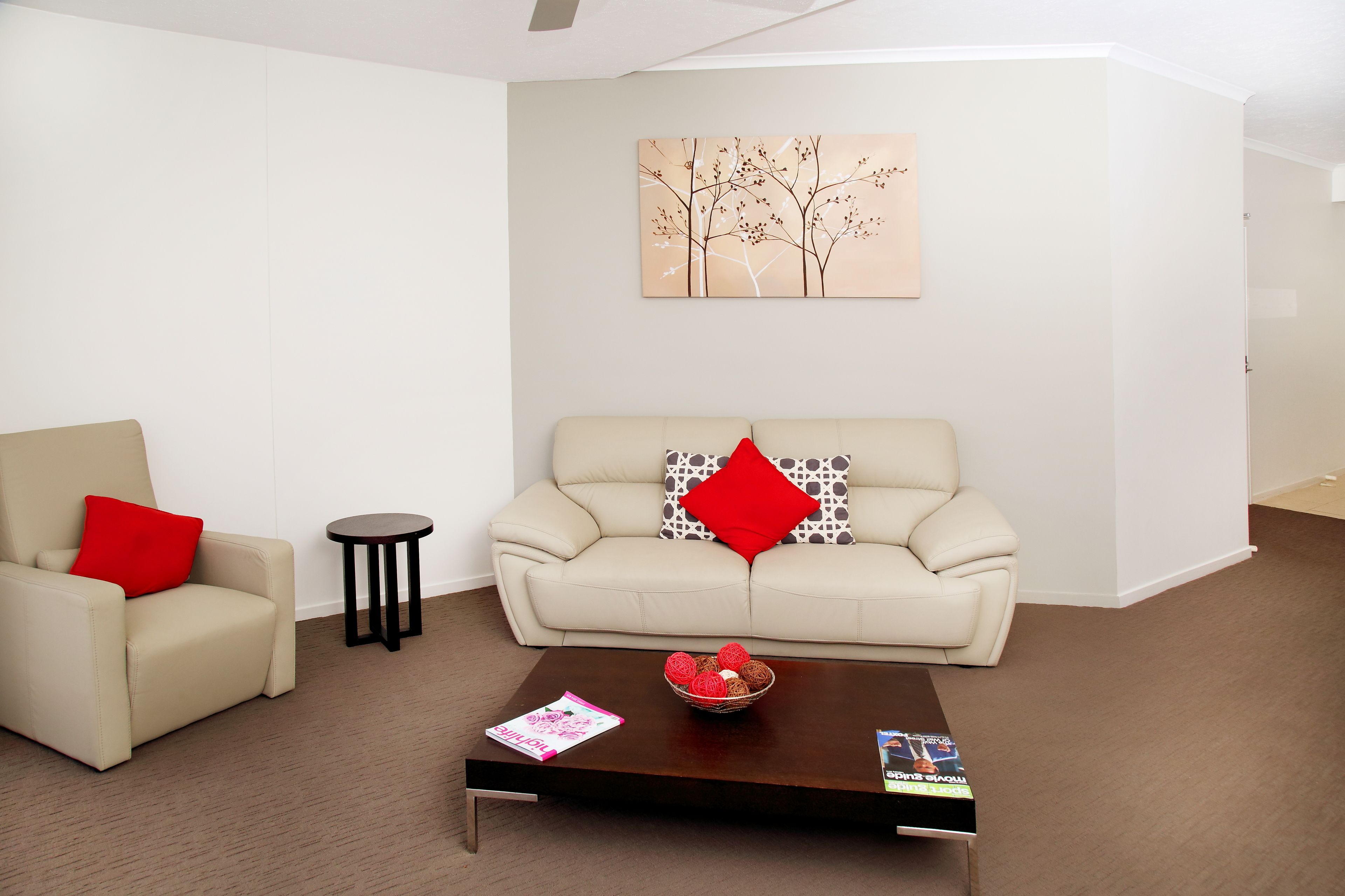 A modern living room with a beige sofa, a red pillow, a small round table, and a coffee table with magazines.