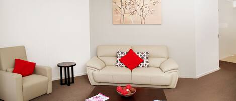 A modern living room with a beige sofa, a red pillow, a small round table, and a coffee table with magazines.