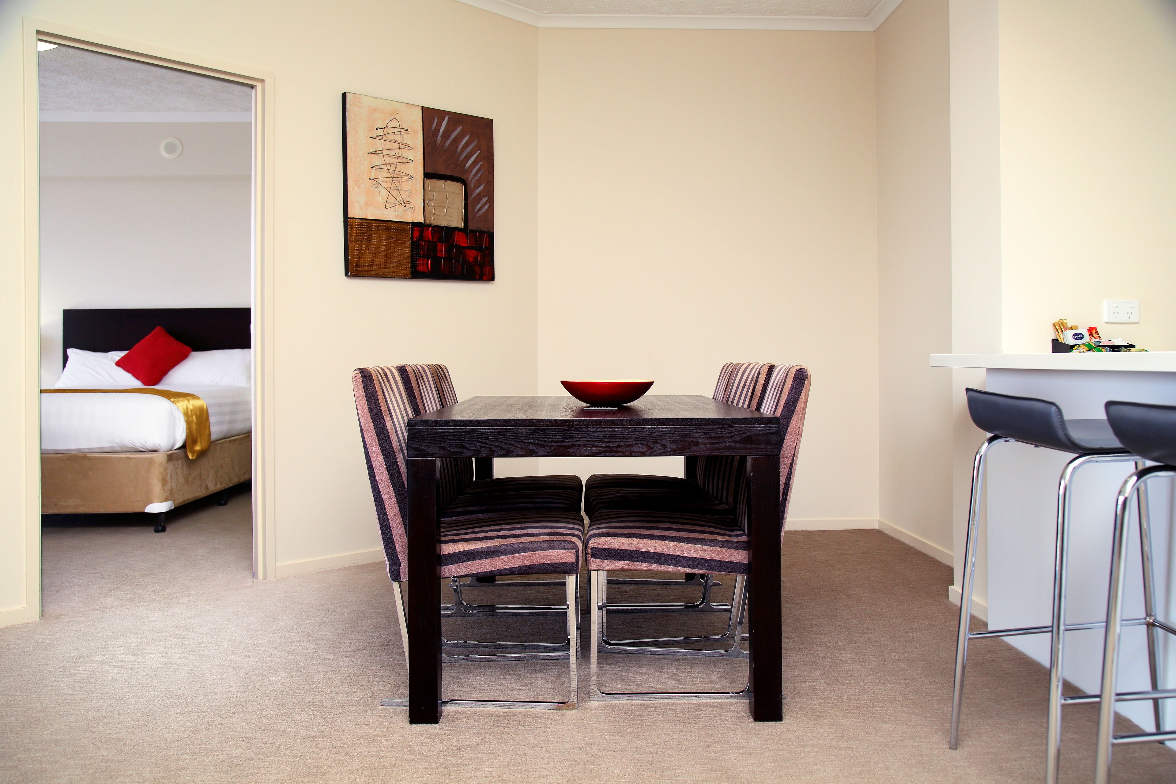 A hotel room with a dining area featuring a wooden table and striped chairs, a bed with a red pillow, and a painting on the wall.