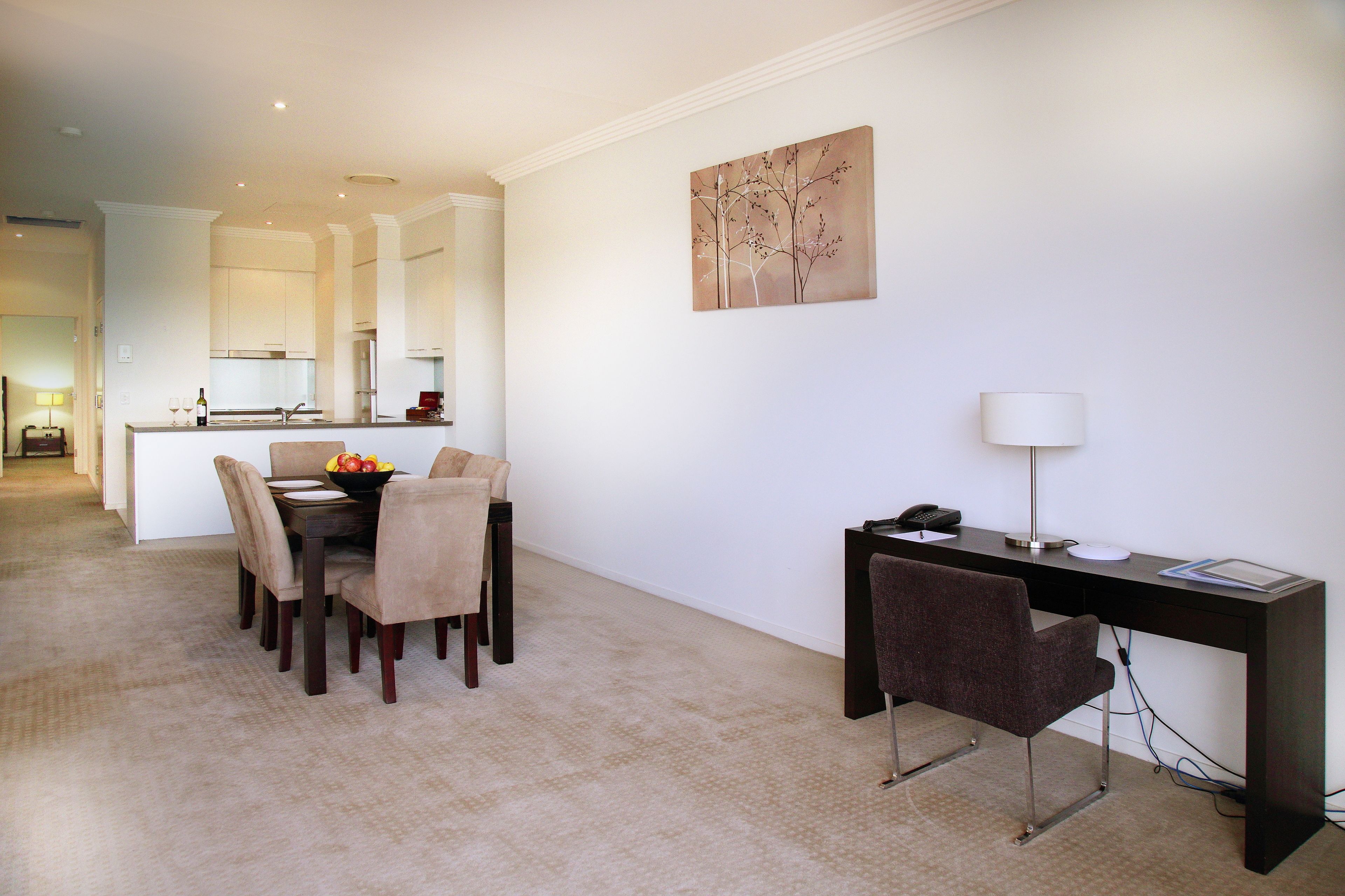 A modern dining area with a table set for four, a dark desk with a lamp, and a painting on the wall.
