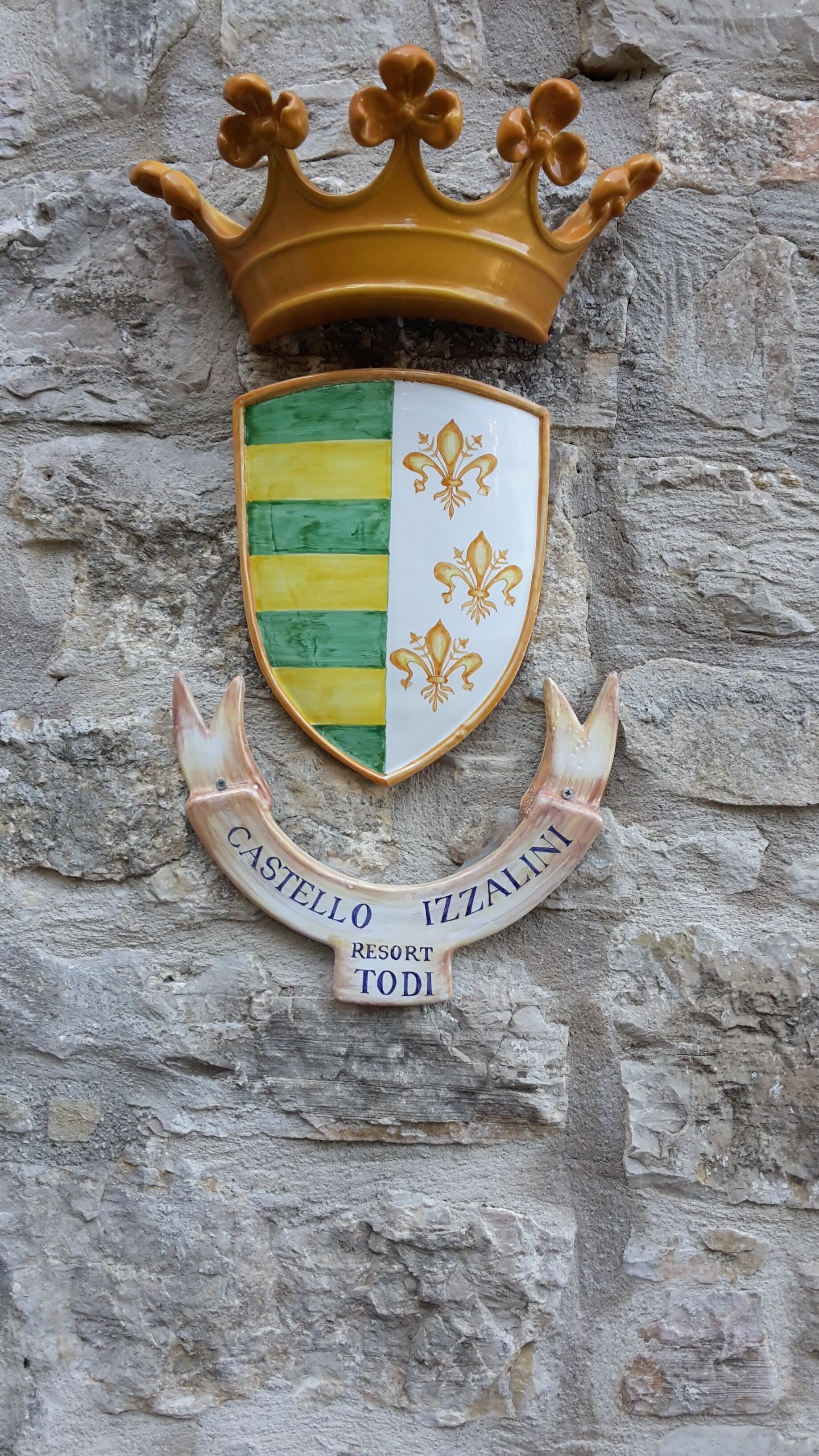 Un blason orné d’une couronne, un écu aux rayures vertes et jaunes, et une bannière portant l’inscription « CASTELLO IZZALDI RESORT TODI ».