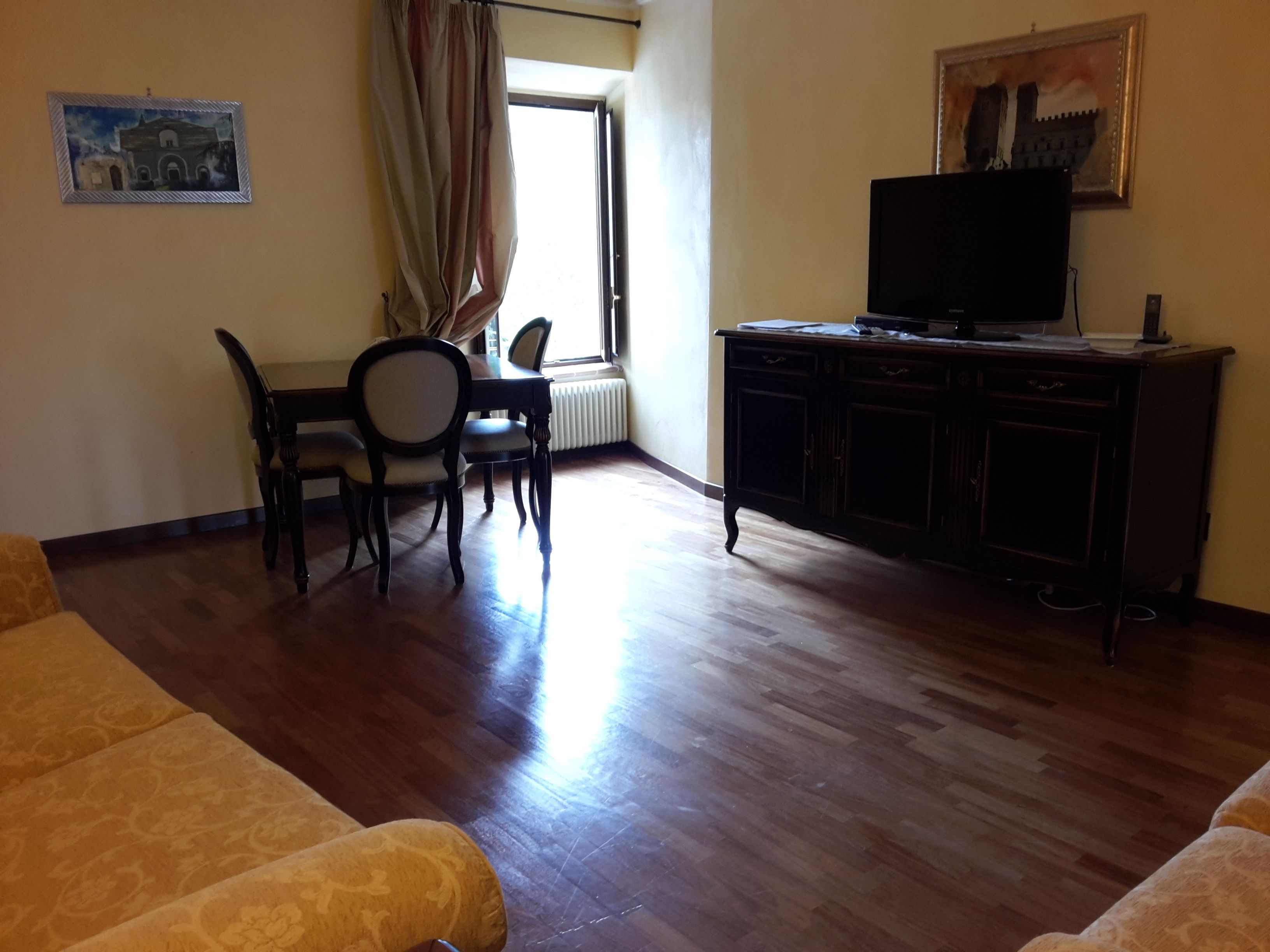 A living room with a sofa, a dining table with chairs, a television on a cabinet, and a window with curtains.