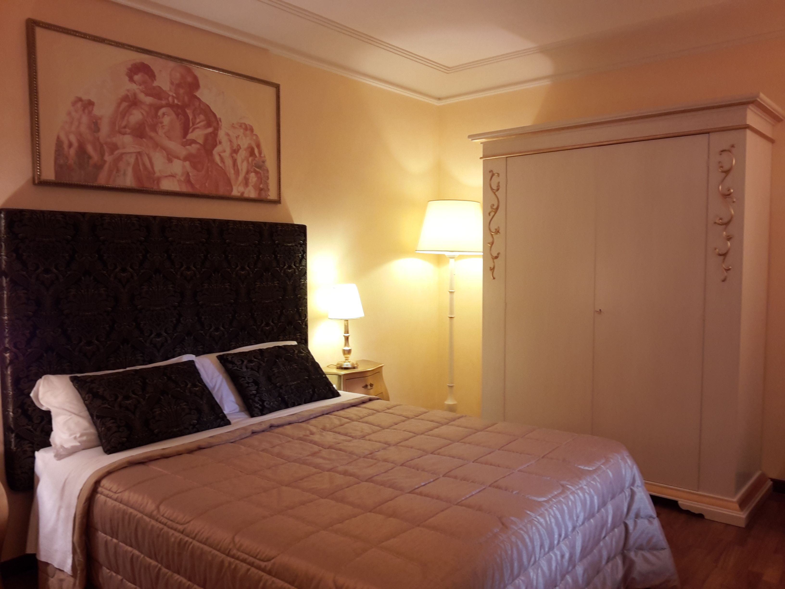 Un dormitorio con una cama grande, cabecera, mesita de noche con lámpara, armario y un cuadro en la pared.