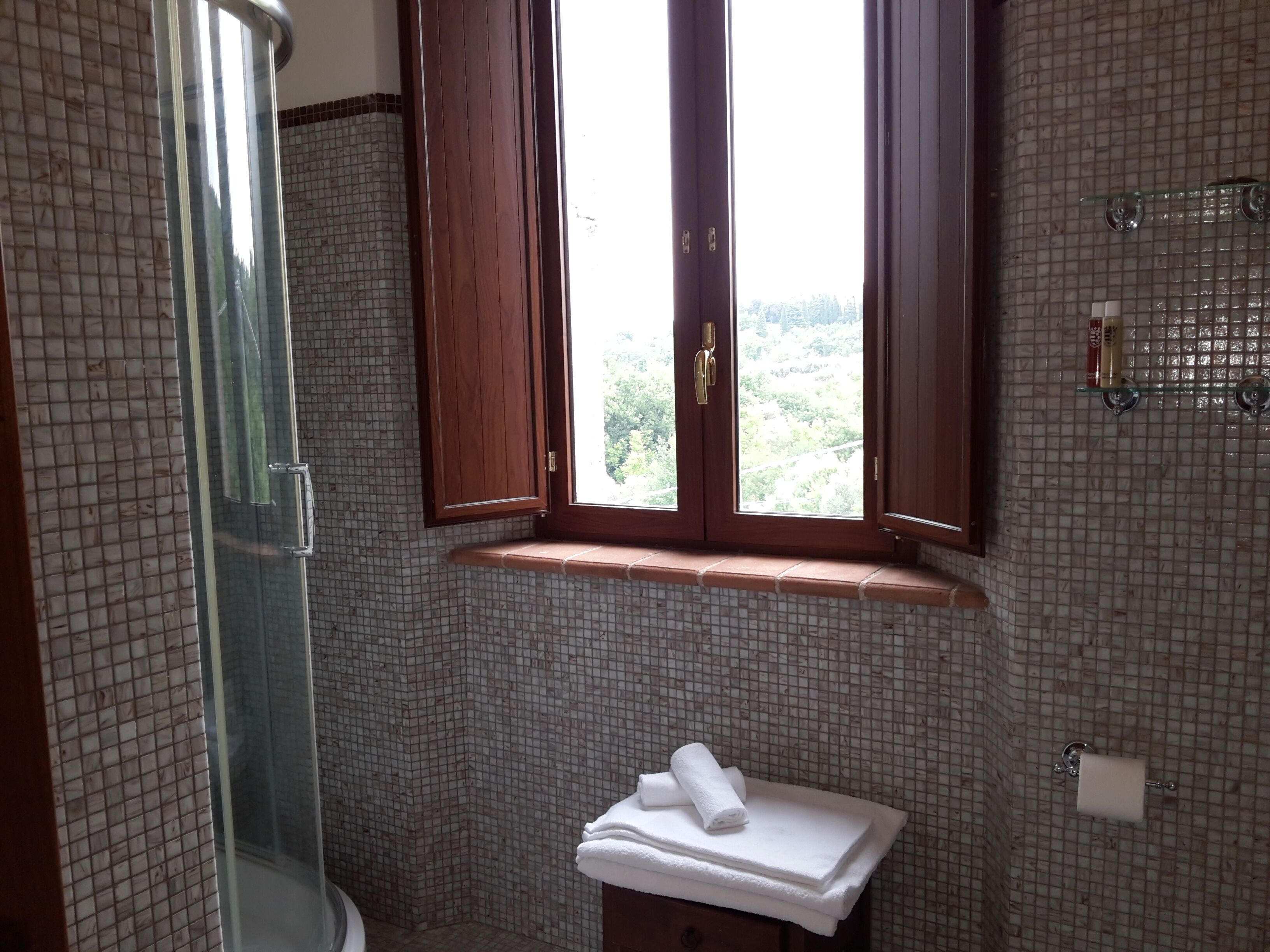 A bathroom with a tiled wall, a window with wooden shutters, a glass shower enclosure, and a towel rack.
