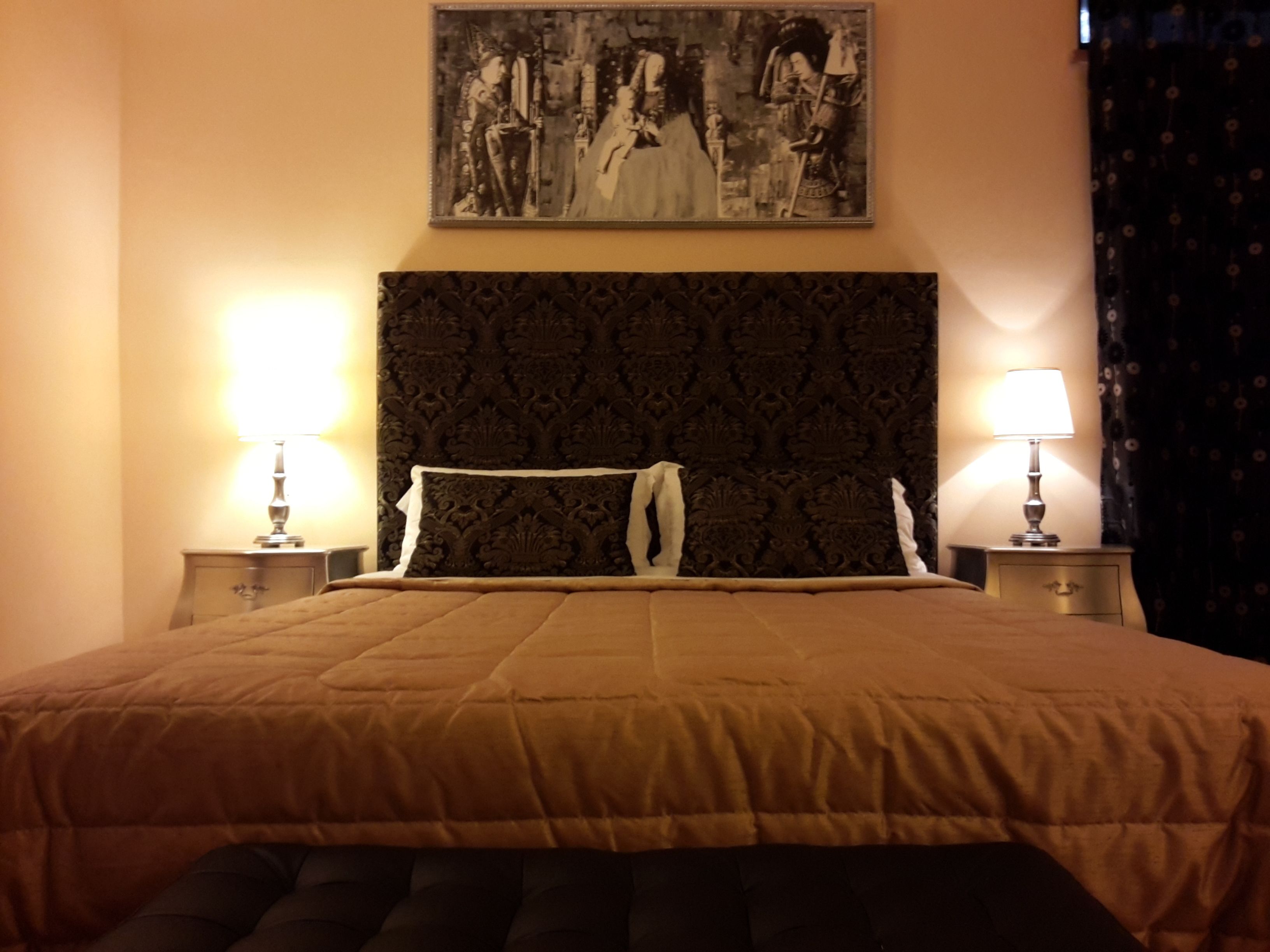 A hotel room with a large bed, two bedside lamps, a decorative headboard, and a framed artwork on the wall.