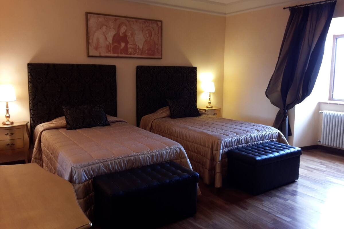 A hotel room with two beds, a large painting on the wall, a window with curtains, and a radiator.
