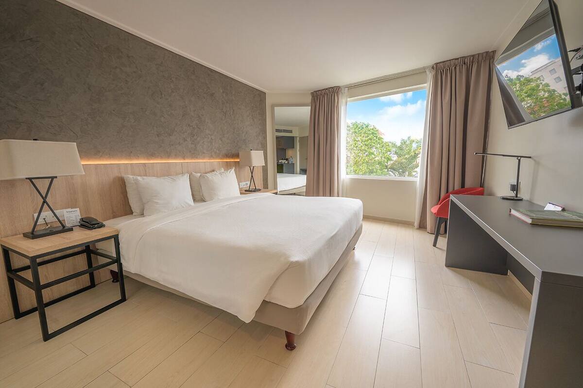 Habitación de hotel con una cama grande, mesitas de noche con lámparas, un escritorio y un ventanal con cortinas.