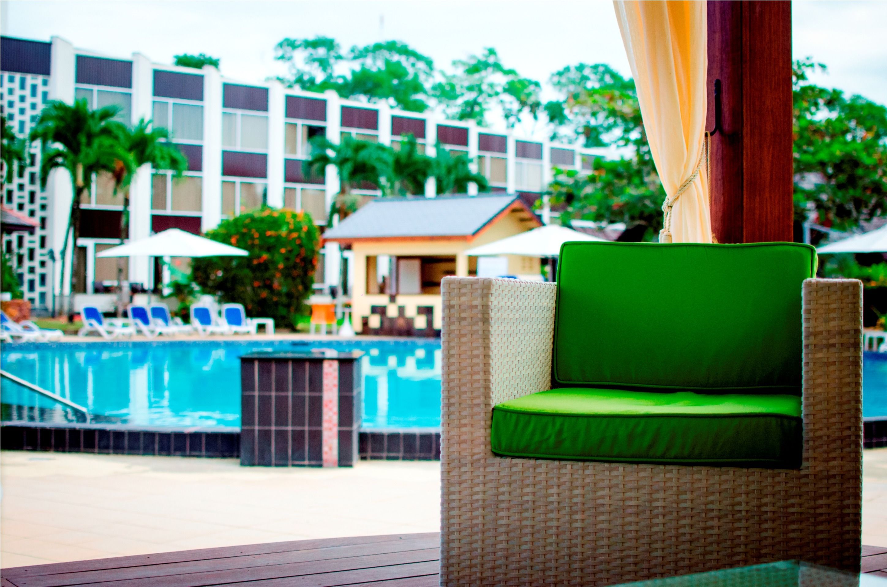 Una silla de mimbre con un cojín verde junto a una piscina, con vista despejada a la piscina, y un edificio con varios balcones al fondo.