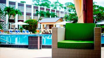 Una silla de mimbre con un cojín verde junto a una piscina, con vista despejada a la piscina, y un edificio con varios balcones al fondo.