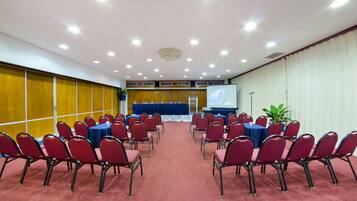 Un salón de conferencias con filas de sillas rojas, un escenario con mantel azul y una pantalla de proyector.