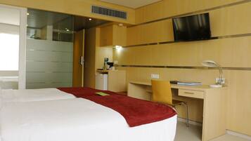 Quarto de hotel com cama, escrivaninha, cadeira, televisão e banheiro com espelho.