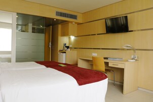 Quarto de hotel com cama, escrivaninha, cadeira, televisão e banheiro com espelho.