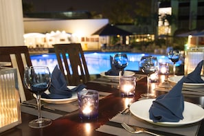 A dining table set with wine glasses, plates, and napkins, with a pool and buildings in the background. - Torarica Resort (Paramaribo)