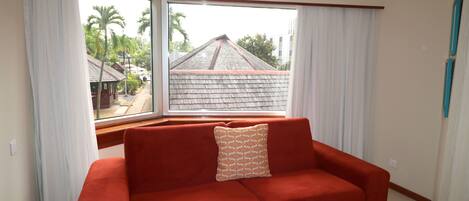A red sofa with a patterned pillow in a room with large windows, white curtains, and a view of a building with a sloped roof.