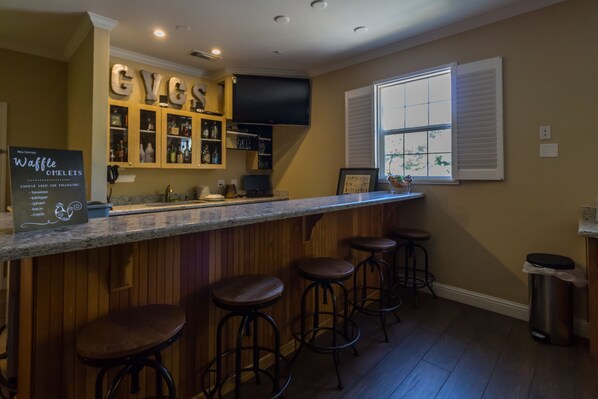 A kitchen with a bar area, wooden bar stools, a sign for waffle omelets, and a window with blinds.