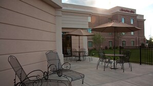 An outdoor patio area with metal chairs and tables, a large umbrella, and a brick building in the background. - Hampton Inn Burlington (Burlington)