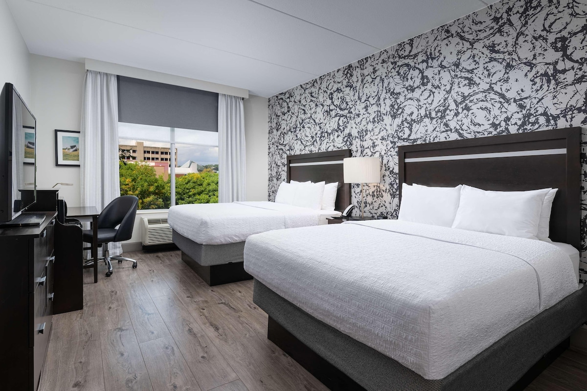 A hotel room with two beds, a desk, a chair, and a large window with a view of greenery and buildings.