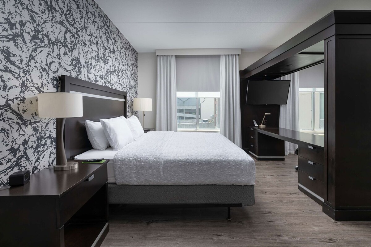 A hotel room with a bed, bedside tables, a TV, and a large window with curtains.
