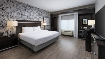 A modern hotel room with a large bed, a desk, and a chair. There is a window with curtains, a wall-mounted TV, and a desk lamp.