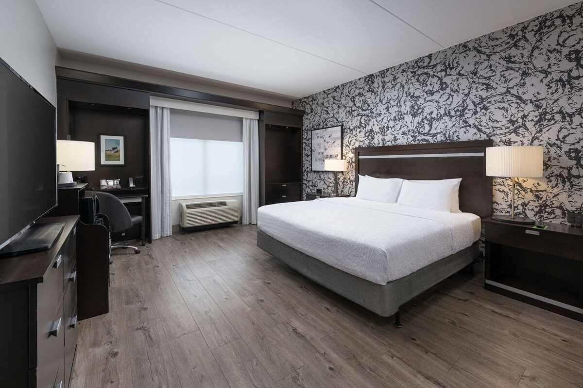 A hotel room with a large bed, a desk, a chair, a TV, and a window with curtains.