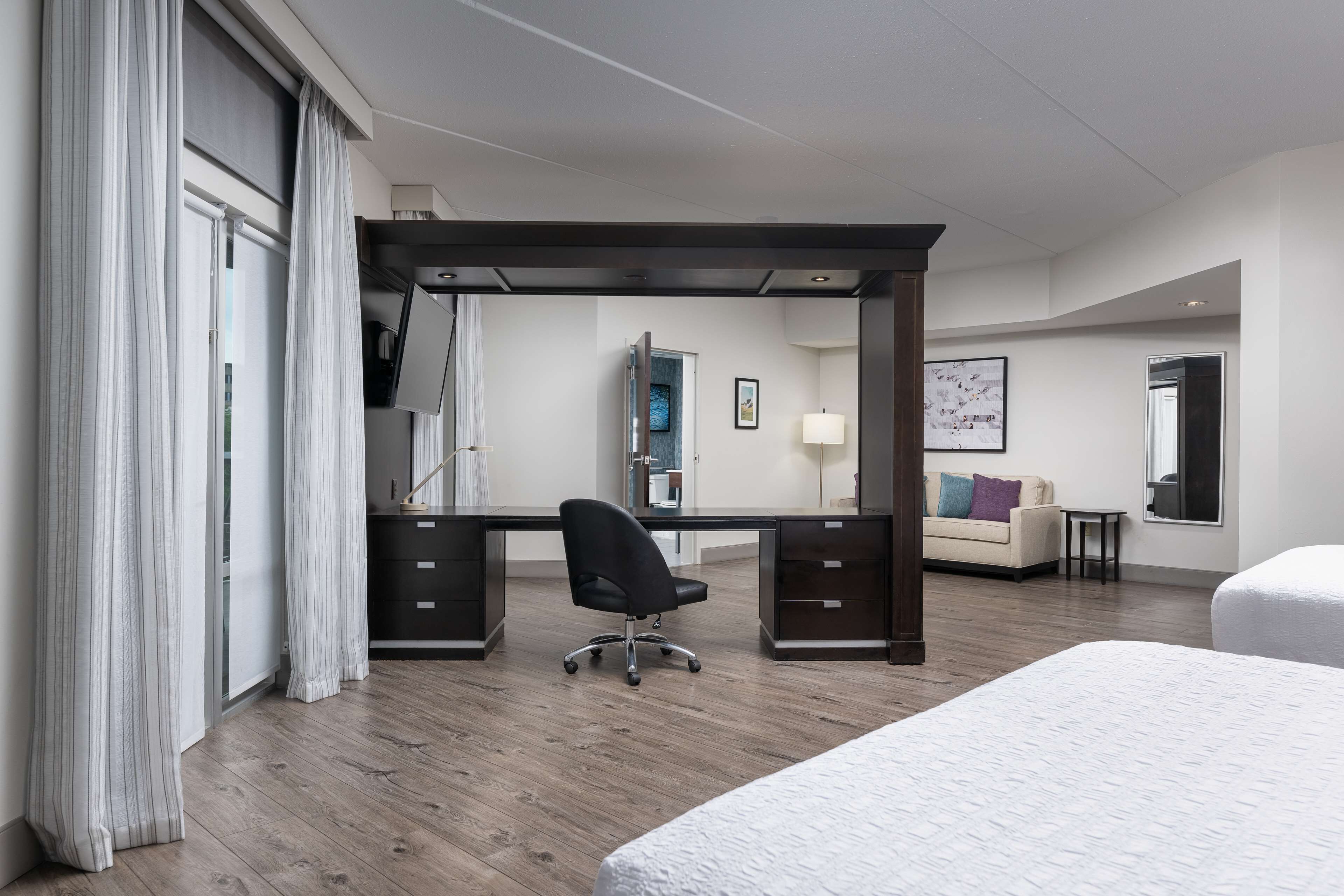 A hotel room with a wooden desk, a black office chair, a bed, a sofa, and a small table.