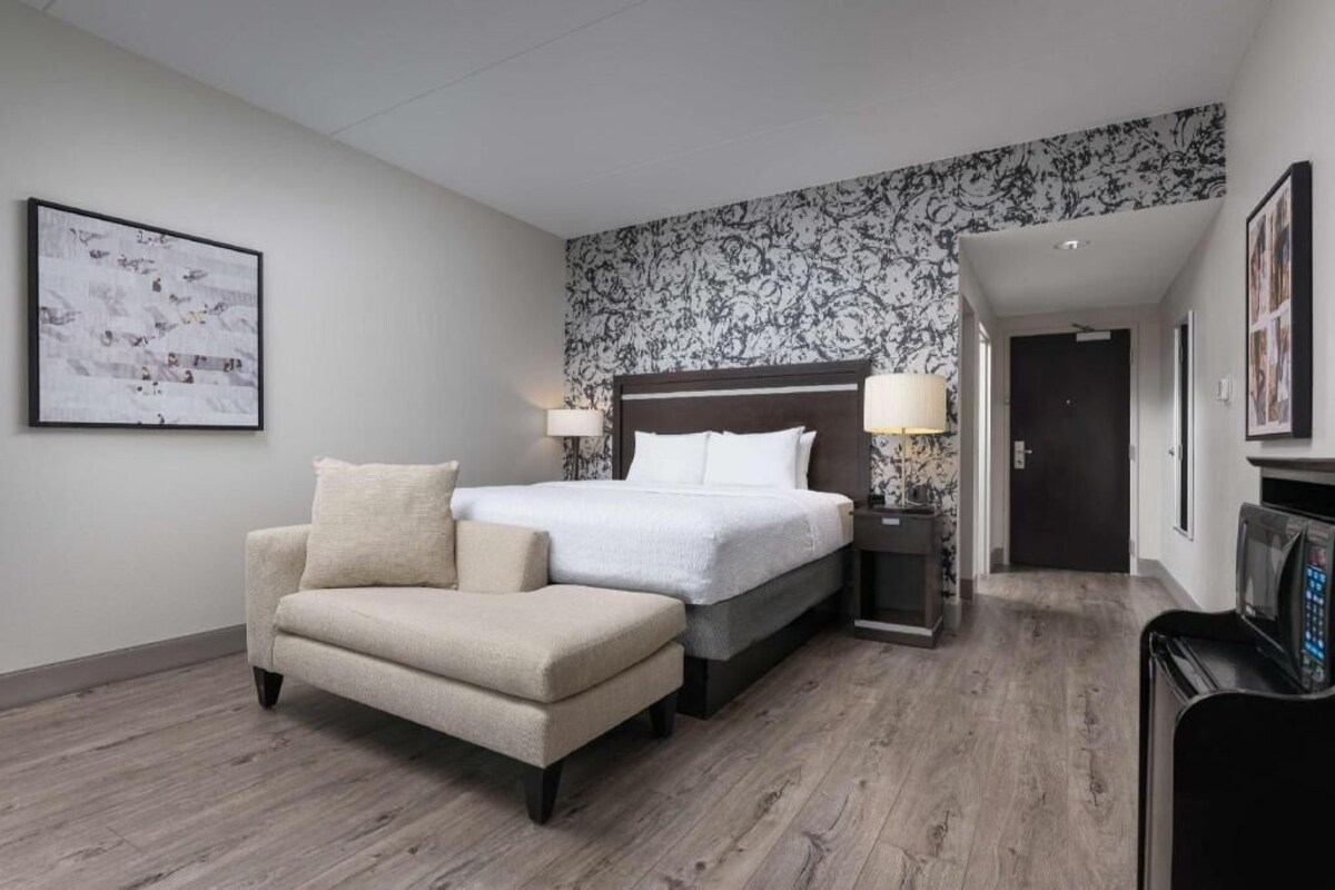 A hotel room with a bed, two bedside lamps, a chair, a TV, and a patterned wall.