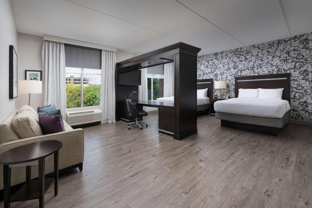 A hotel room with a bed, a desk, a chair, a sofa, and a window with curtains.
