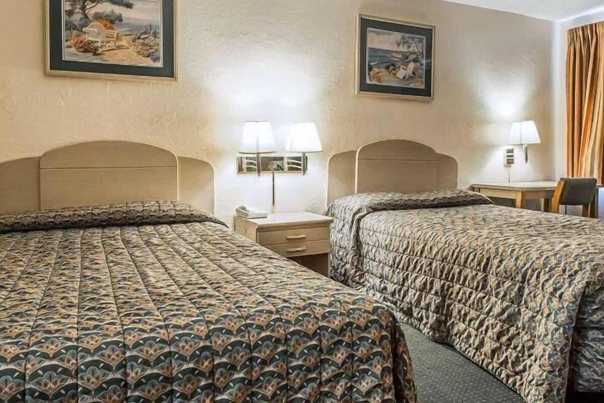 Quarto de hotel com duas camas, uma mesinha de cabeceira, um abajur e uma cadeira.
