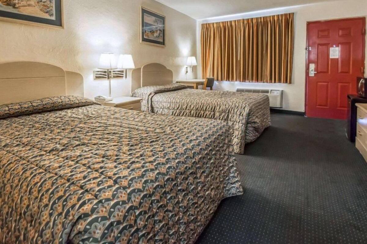 Quarto de hotel com duas camas, uma porta vermelha, janela com cortinas e um quadro na parede.